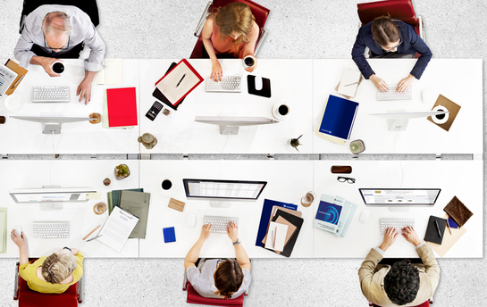 Sechs Personen sitzen an einem langen Tisch mit Computern und Arbeitsmaterialien, von oben betrachtet.