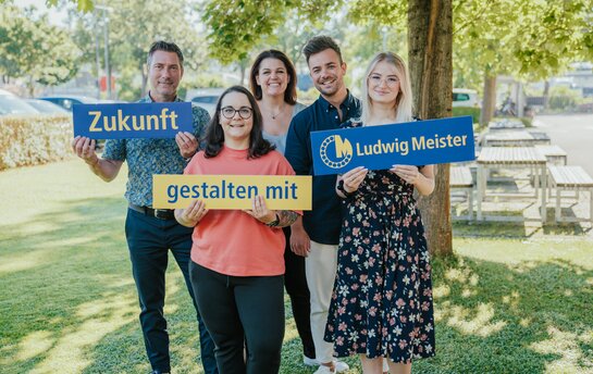 Eine Gruppe von fünf Personen steht draußen auf einer Wiese und hält Schilder mit den Worten "Zukunft gestalten mit Ludwig Meister".