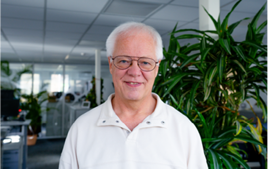 Ein älterer Mann mit Brille und weißem Hemd steht in einem Büro vor einer großen Zimmerpflanze.