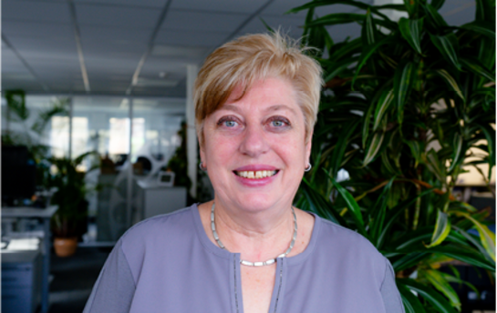 Eine Frau mit kurzem blondem Haar lächelt in einem Büro mit großen Pflanzen im Hintergrund.