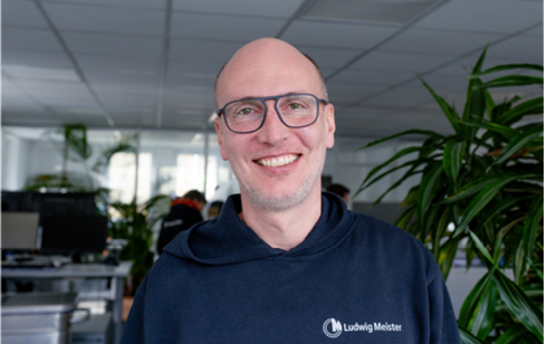 Ein Mann mit Brille und dunklem Hoodie steht lächelnd in einem modern eingerichteten Büro.