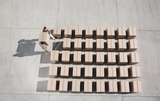 Zwei Personen tragen Kartons, die in mehreren Reihen auf einem Betonboden im Freien gestapelt sind.