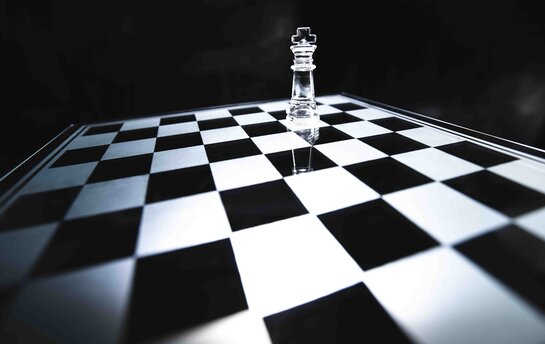 Eine einzelne Glas-Schachfigur steht auf einem Schachbrett in einem dunklen Raum.