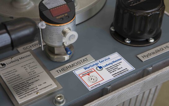 Nahaufnahme eines Hydrauliksystems mit einem Thermostat und einer Wartungsplakette der Firma Ludwig Meister.