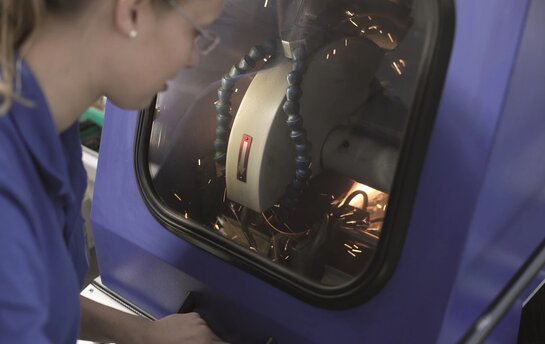 Eine Person arbeitet an einer Maschine, die Funken erzeugt, durch ein Fenster in einem Schutzgehäuse.