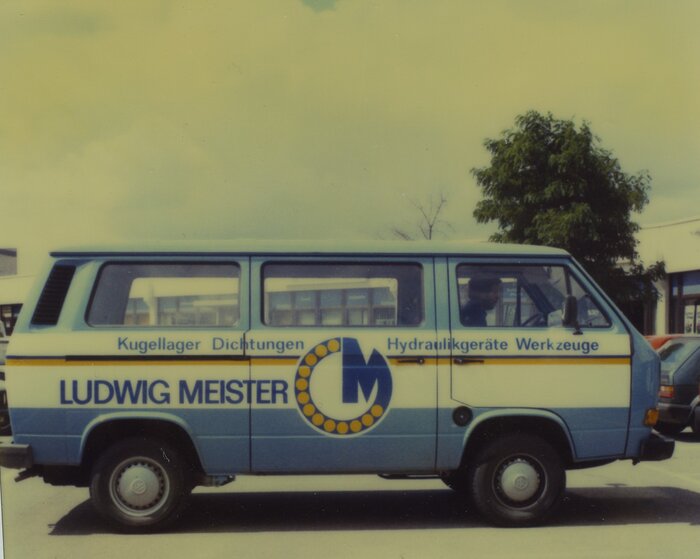 Ein blauer VW-Bus mit der Aufschrift "Ludwig Meister" und Firmenschriftzug, geparkt auf einem Parkplatz vor einem Baum und Gebäuden.