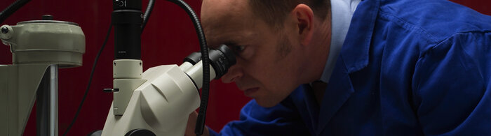 Eine Person in blauer Arbeitskleidung blickt durch ein Mikroskop.