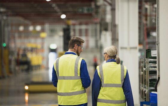 Zwei Personen in Arbeitskleidung gehen durch eine Industriehalle.